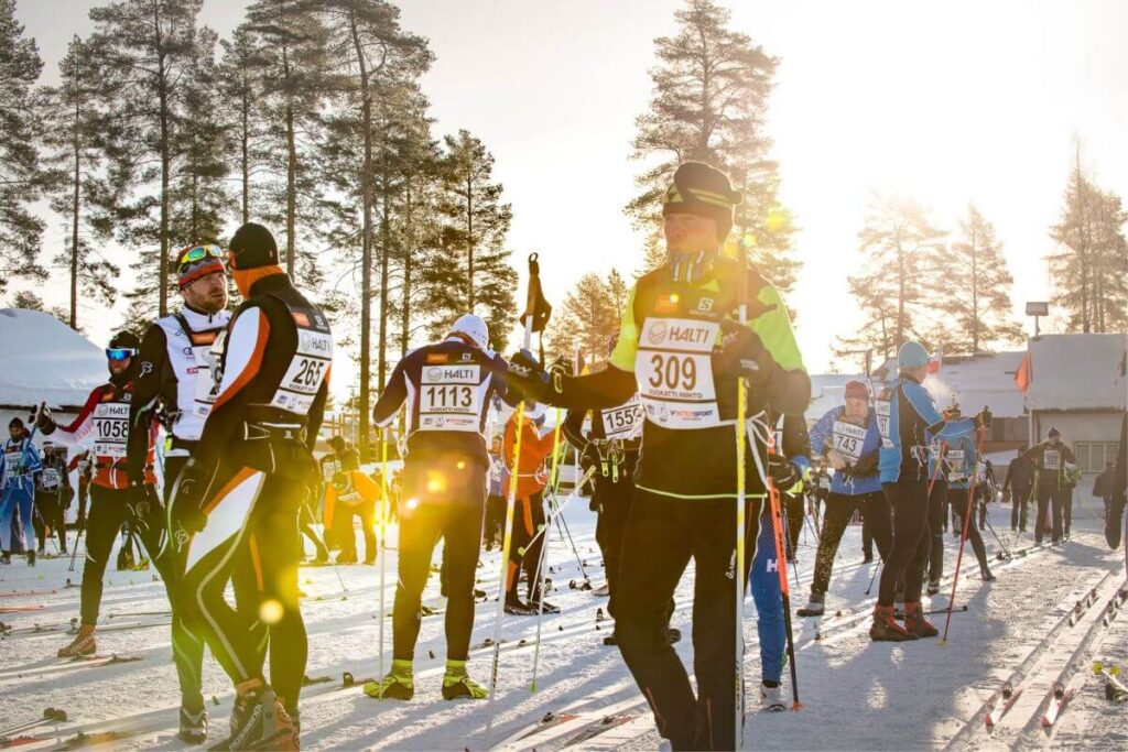 Monia hiihtäjiä hiihtoladulla, aurinko taustalla.