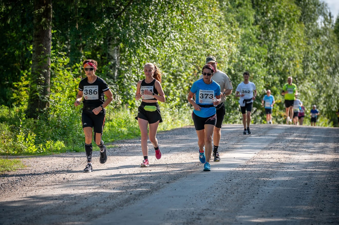 Henkilöt juoksevat hiekkatiellä. Taustalla puita.